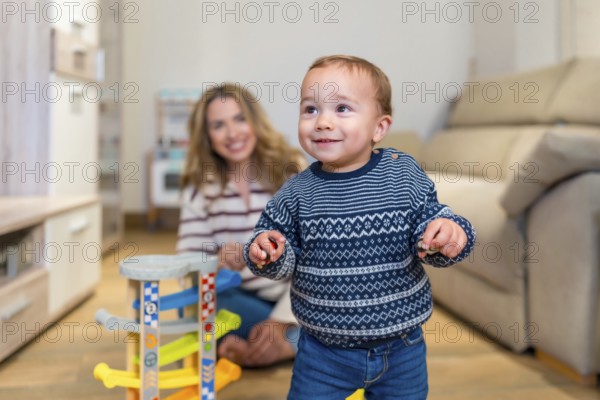Happy baby boy learning to walk in living room with his mother watching and smiling, playing with a