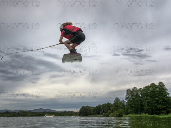 Boy or girl flies over the lake, jump with wakeboard, red life jacket, water skiing in wakepark,