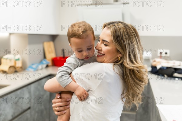 Smiling mother holding her baby son in a home kitchen, enjoying a tender moment of affection and