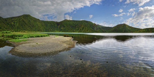 Calm lake surrounded by green mountains under a partly cloudy sky, crater lake Lagoa do Fogo,