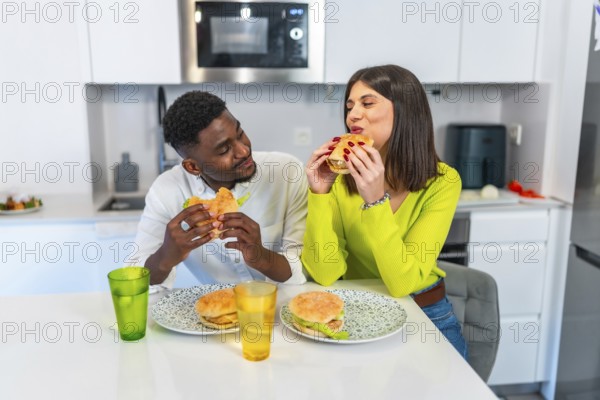 Young multiethnic couple eating burgers in their modern kitchen, enjoying a meal together