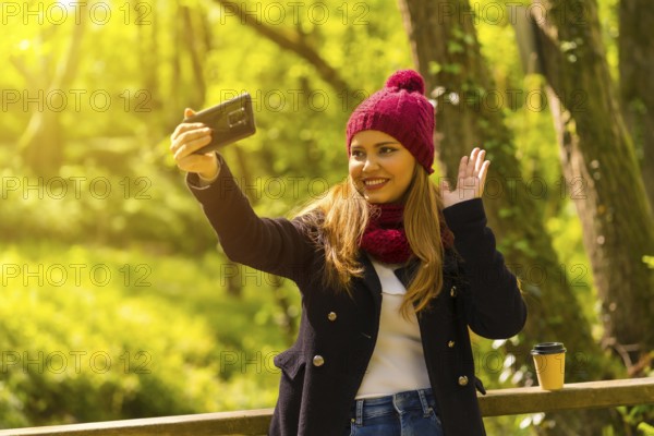Young man in a black jacket, scarf and red woolen hat enjoying in an autumn park, waving on a video