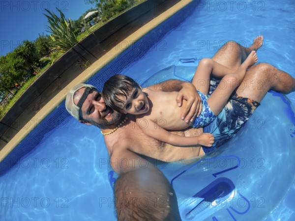 Father and son are taking a selfie while relaxing on an inflatable ring in a swimming pool at a
