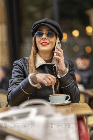 Fashionable woman wearing sunglasses and hat stirring coffee while talking on mobile phone at a