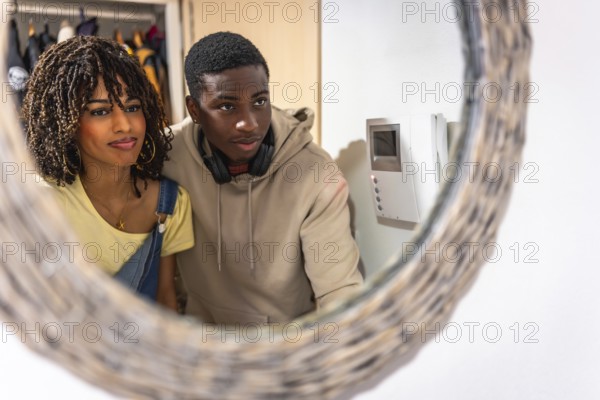 Young multiethnic couple looking at intercom screen reflected in a mirror in the entrance hall of