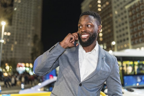 Young businessman smiling and chatting on a smartphone in a vibrant urban city at night, surrounded