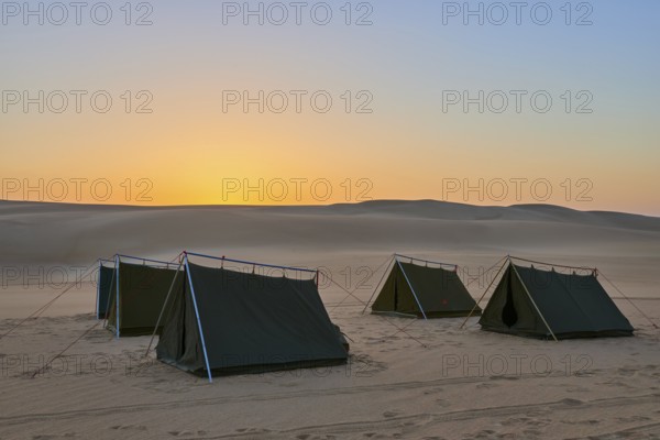 Four tents stand in a vast desert at dusk, surrounded by sand dunes under an orange-coloured sky,