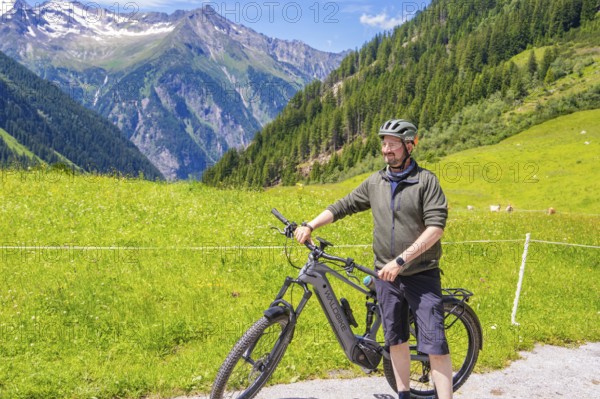 Man in sportswear standing next to his bicycle on a green meadow, surrounded by mountains and blue
