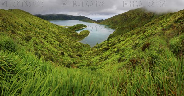 Natural landscape with a green slope and a lake surrounded by hills and a cloudy sky, crater lake