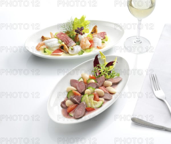 Two plates each with a meat dish and a fish dish, a glass with white wine, a fork on a napkin,