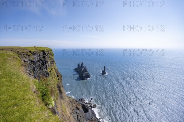 Young man standing on cliff, view from steep cliff to the rocks Reynisdrangar in the water, at