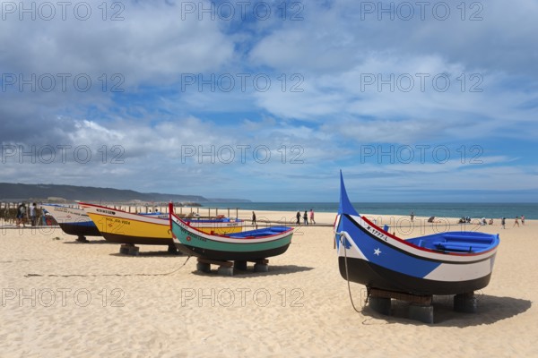 A row of colourful fishing boats on the sandy beach, some people walking along the shore, Praia da