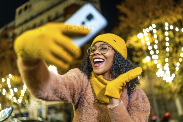 Smiling woman wearing yellow woolen gloves, hat and scarf taking selfie with her smartphone in a