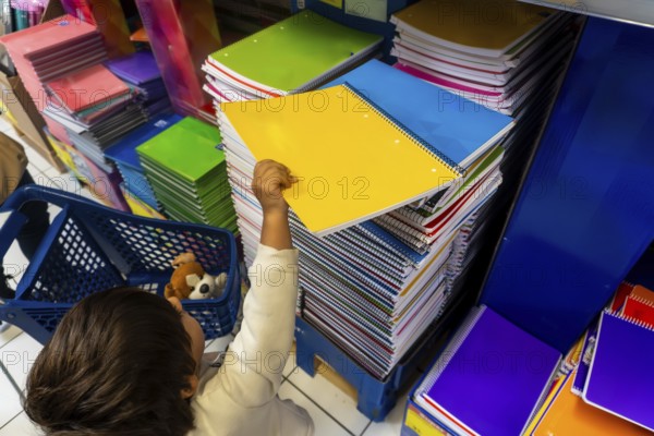 A child is reaching for a yellow spiral notebook in a store. The store is filled with many