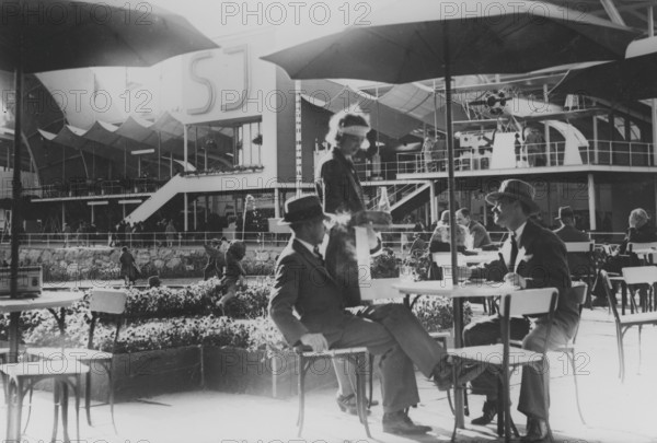 1930s open-air cafe. Two men are being served drinks at the table by a waitress on the grounds of