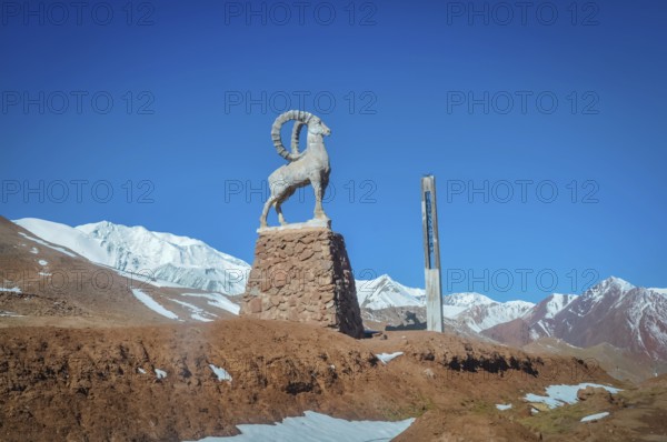 Sculpture of bouquetin on stony base surrounded by mountains with snowy peaks in distance in