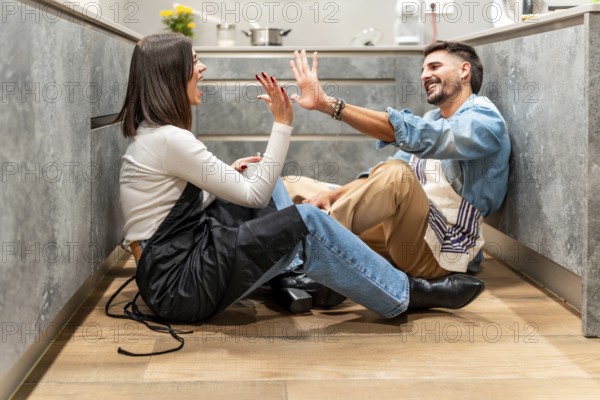 Two young chefs are sitting on the kitchen floor, giving each other a high five and laughing,