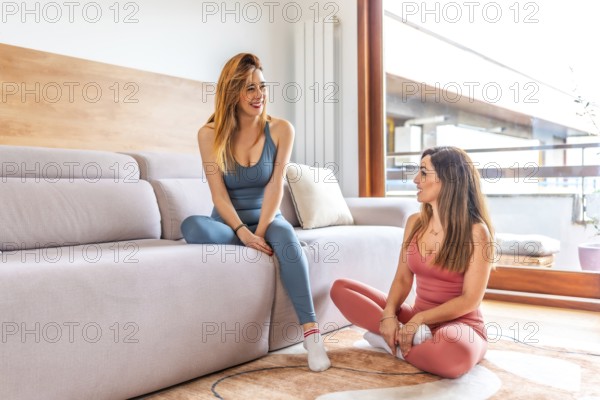 Two smiling women wearing yoga clothes are chatting and relaxing in a modern living room after