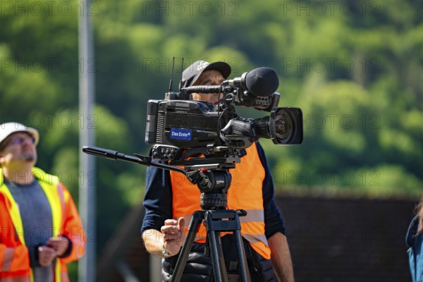 A cameraman in a brightly coloured waistcoat with a camera films outside in sunny weather against a