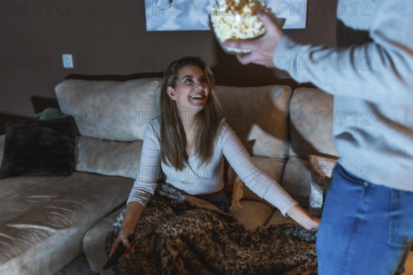 Smiling woman sitting on sofa with remote control, covered with blanket, taking popcorn from