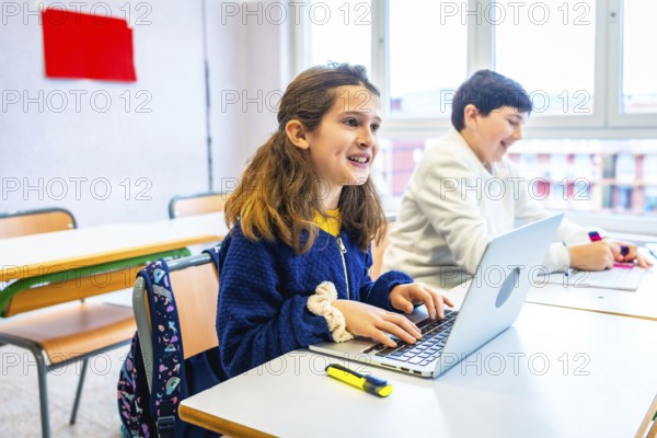 Smiling elementary school girl using laptop during lesson in classroom, next to classmate writing