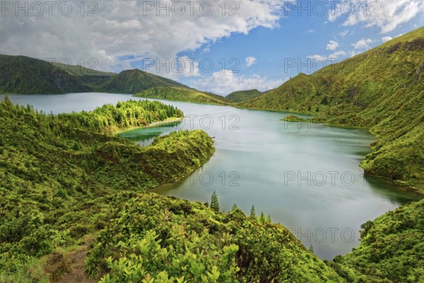 A tranquil lake surrounded by green mountains and wooded slopes under a blue sky, crater lake Lagoa