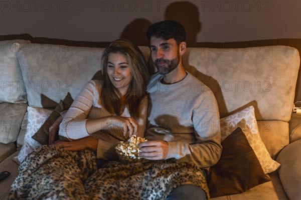 Young couple enjoying a movie night at home, sharing popcorn and relaxing on the sofa under a cozy
