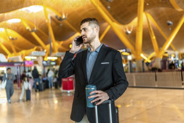 Businessman making a phone call while holding a reusable coffee cup and pulling his luggage at a