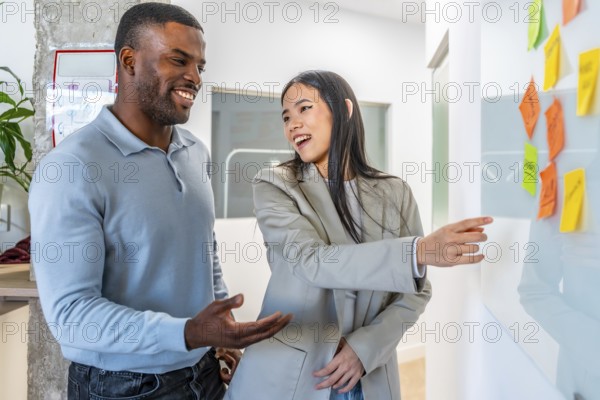 Two smiling businesspeople discussing work related tasks pointing at sticky notes on whiteboard in