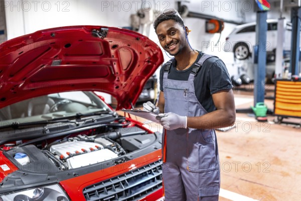 Smiling mechanic taking notes on clipboard while inspecting engine of red car with open hood in a