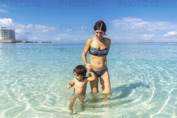 Mother and son holding hands and wading in the crystal clear turquoise waters of estanys beach in