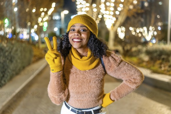Smiling woman wearing yellow winter clothes showing victory sign while enjoying christmas lights in