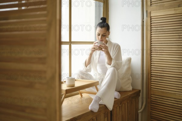 Pretty woman enjoys a peaceful morning, sipping coffee while sitting on windowsill, basking in the