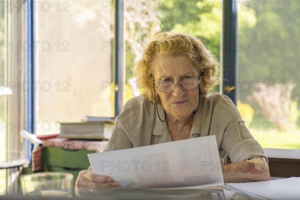 Focused elderly woman with glasses reviewing paperwork, possibly bills or medical records, sitting