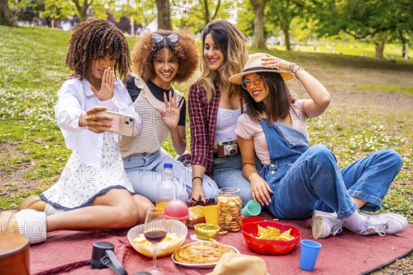Four cheerful young women enjoying a picnic in a park, taking a joyful selfie with a smartphone