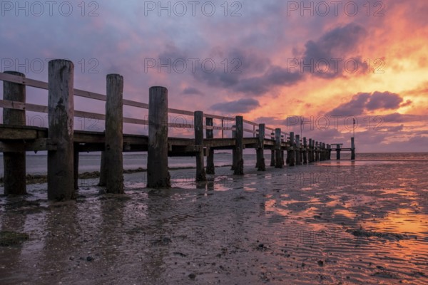 A jetty on the beach at sunset, the sky is coloured orange and purple, the water reflects the