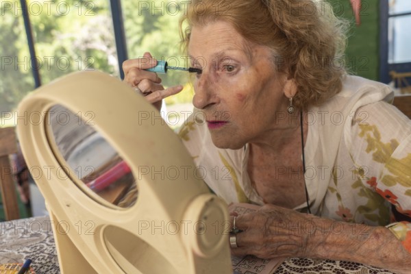 Elderly woman applying mascara with care, using a small mirror on the table, enhancing her beauty