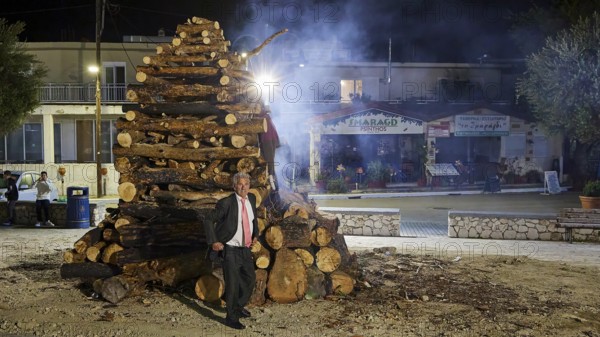 A man in a suit stands at night next to a smoking pile of wood, Mayor of Psinthos, Easter fire