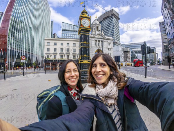 Two happy tourists taking a selfie in london near a historical clock tower, with modern buildings