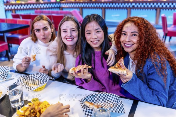 Four diverse young women enjoying a meal together at a restaurant, holding slices of pizza and