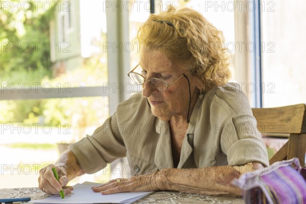 Concentrated elderly woman wearing glasses drawing with green colored pencil on sheet of paper