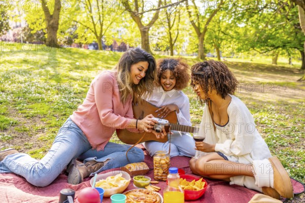 Three friends enjoying a picnic in a park, taking photos with a vintage camera, playing guitar, and