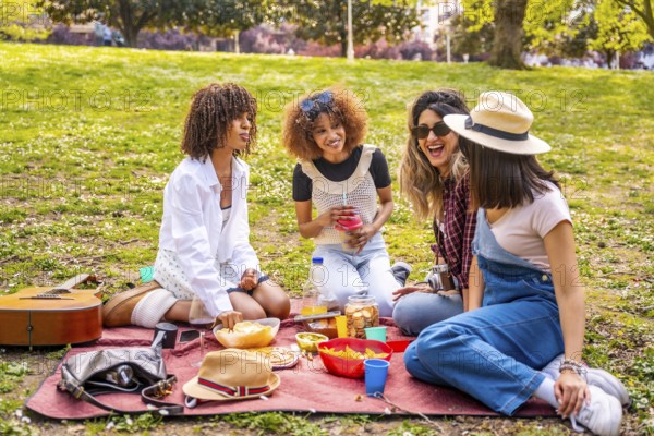 Four diverse female friends enjoying a picnic in a park, sharing food, drinks, and laughter on a