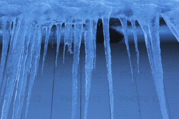Long icicles and thick heavy ice hanging from roof edge of grey wood plank cladded building in
