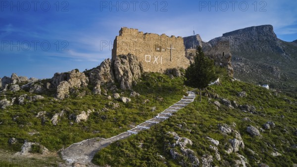 Drone shot, stone ruin on hill with paved path in mountainous landscape under blue sky,