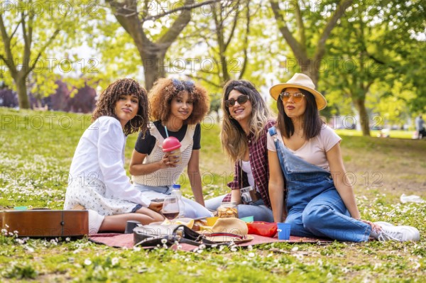 Four diverse female friends enjoying a picnic in a park, sharing food, drinks, and good times on a