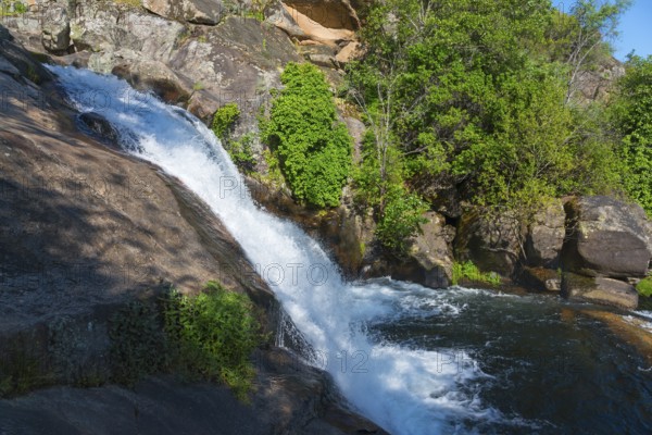 A small waterfall flows over large rocks, surrounded by green plants under a sunny sky, Cascada del