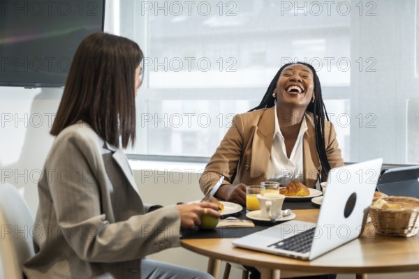 Two businesswomen are enjoying breakfast and laughing together during their business trip, creating