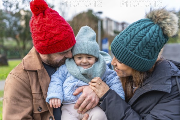 Parents wearing winter hats embracing their smiling baby in a park, enjoying a sunny winter day
