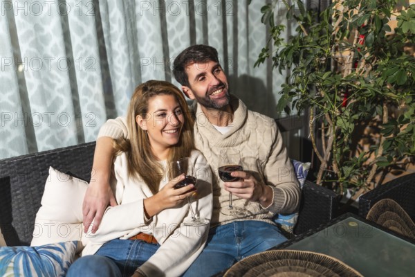 Happy couple relaxing on a comfortable sofa on the terrace, watching a movie and drinking red wine,